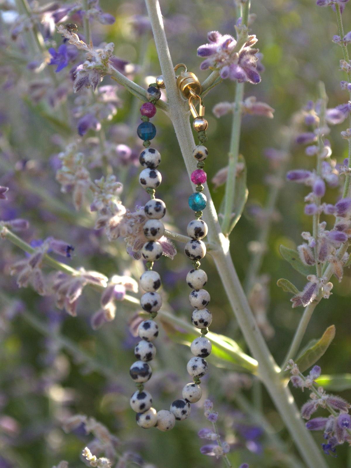 A beaded bracelet with white and black spotted beads, pink beads, and blue-green beads, hanging from a plant stem surrounded by purple flowers.