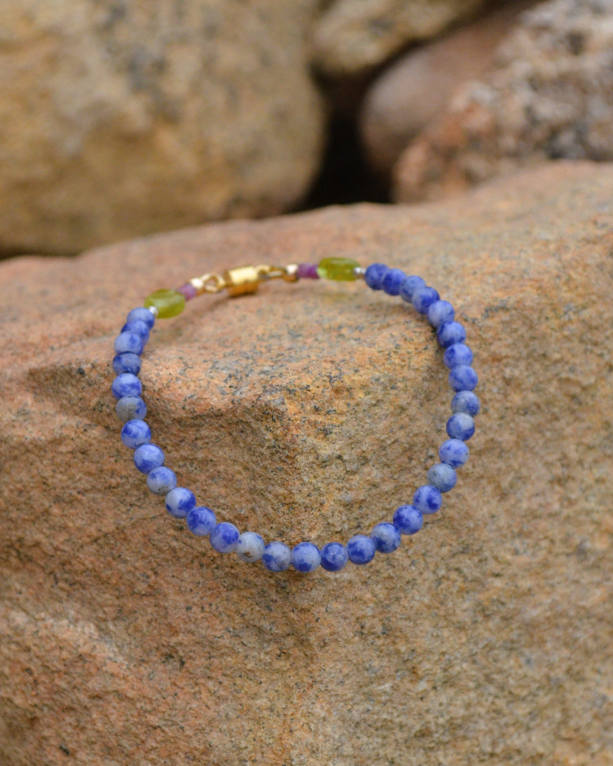 A beaded bracelet made of blue and white marbled beads, accented with small green beads and a gold clasp, resting on a textured brown rock.