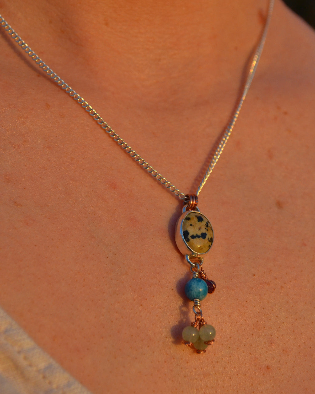 A close-up of a delicate silver chain necklace worn on a person's neck, featuring an oval pendant with a yellow and black marbled pattern, accented by two dangling blue beads and small green stone clusters.