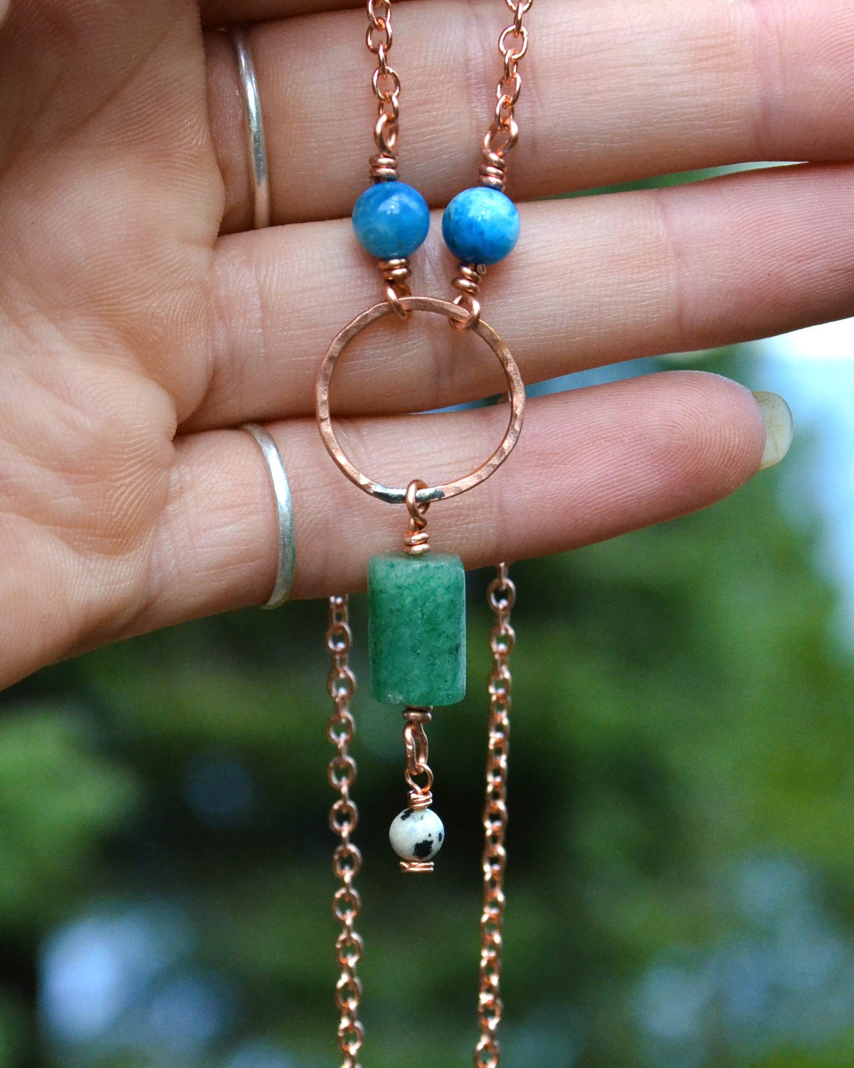 A close-up of a hand holding a handmade necklace with a copper chain, blue beads, a hammered copper ring, a green rectangular stone, and a small white bead charm, against a blurred green outdoor background.