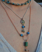 A close-up of a person wearing a layered necklace set. The set includes a silver chain with a round stone pendant, a copper chain with blue beads and a green rectangular bead drop, and a beaded choker.