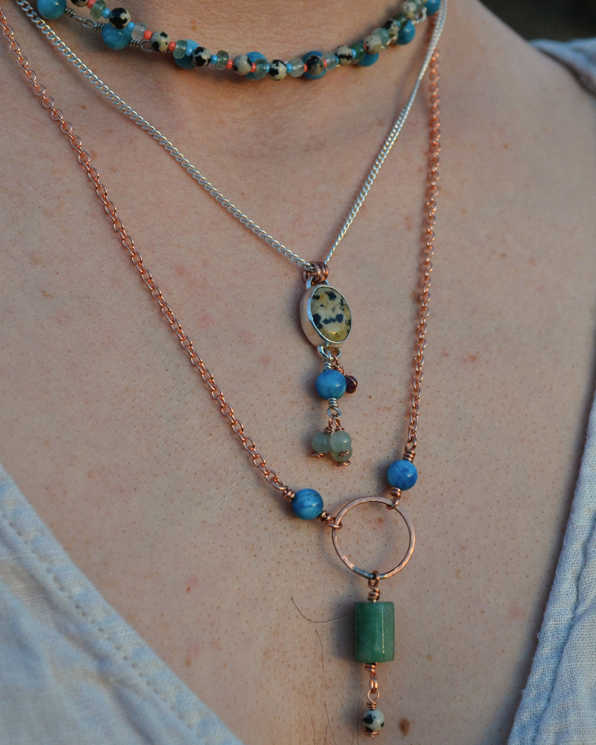 A close-up of a person wearing a layered necklace set. The set includes a silver chain with a round stone pendant, a copper chain with blue beads and a green rectangular bead drop, and a beaded choker.
