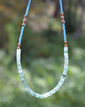 A beaded necklace with light blue and pale green faceted beads, accented with small brown and gray beads, shown against a blurred green outdoor background.