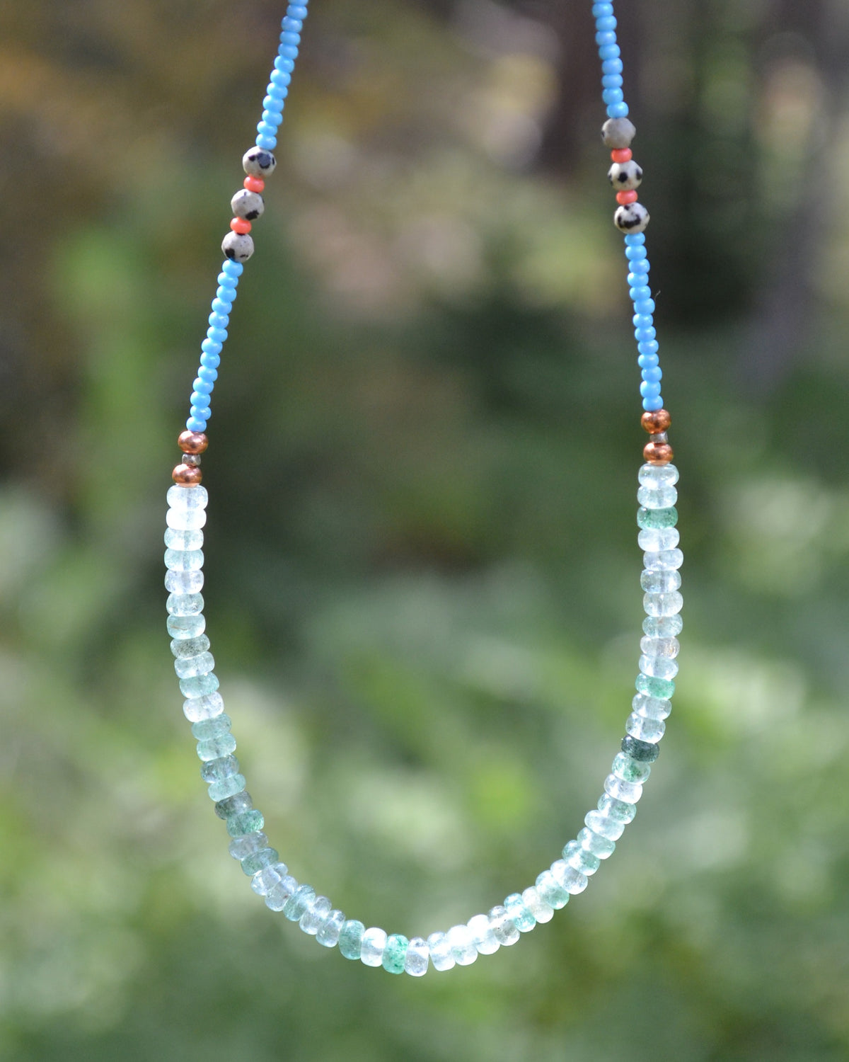 A beaded necklace with light blue and pale green faceted beads, accented with small brown and gray beads, shown against a blurred green outdoor background.