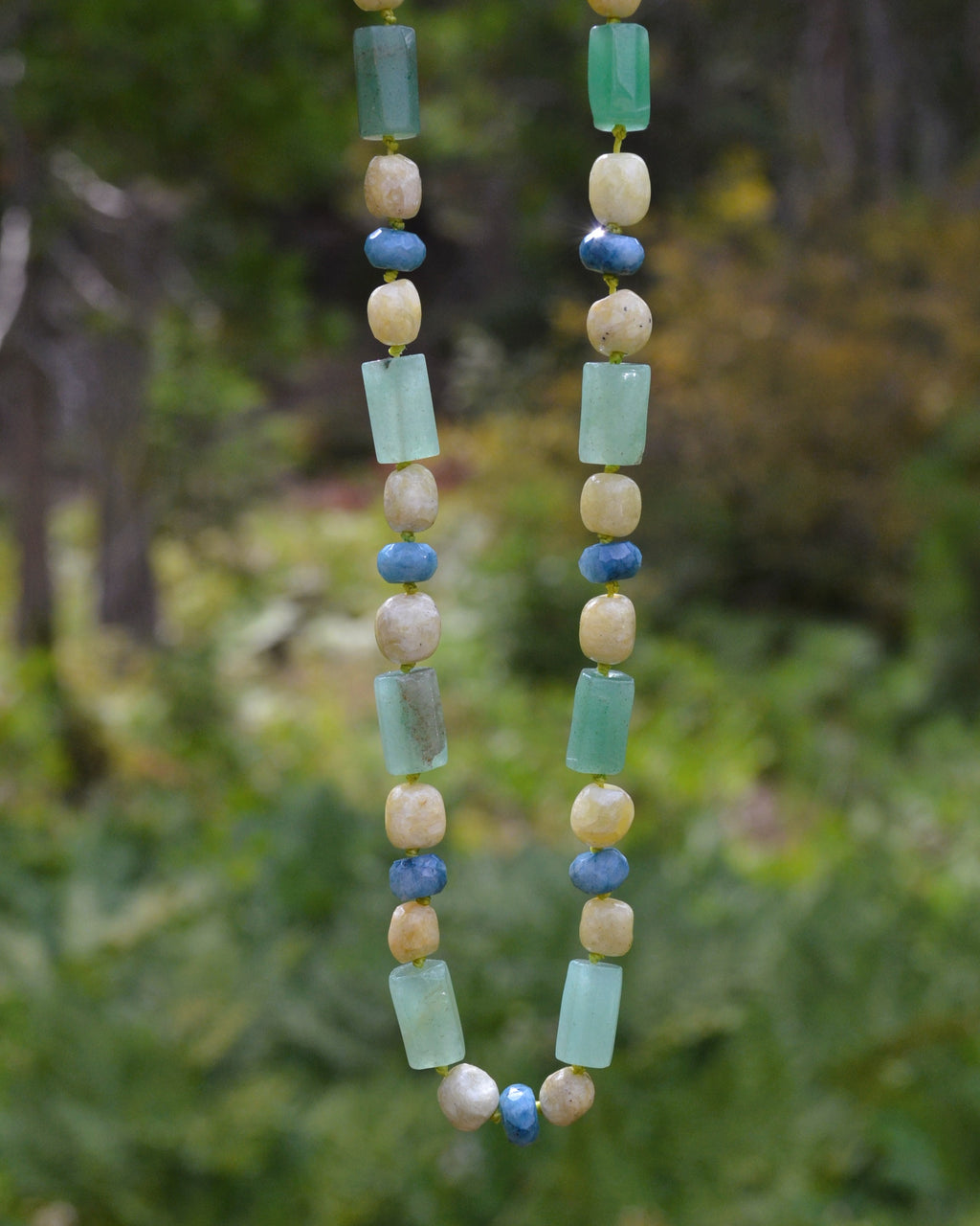 A beaded necklace with light green rectangular beads, cream-colored round beads, and small blue round beads, hanging against a blurred green outdoor background.
