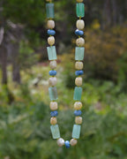 A beaded necklace with light green rectangular beads, cream-colored round beads, and small blue round beads, hanging against a blurred green outdoor background.