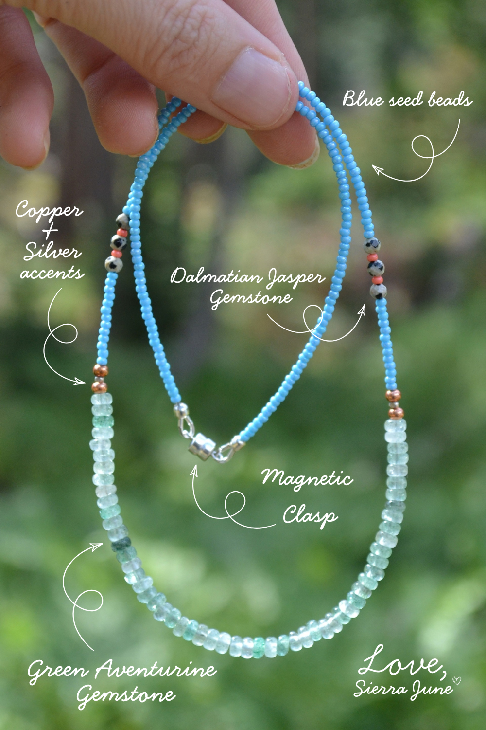 A hand holds a beaded necklace outdoors with a blurred green background. White callout lines and text label the necklace components, including blue seed beads, a Dalmatian Jasper gemstone, copper and silver accents, a magnetic clasp, green aventurine gemstone, and a Love, Sierra June design.