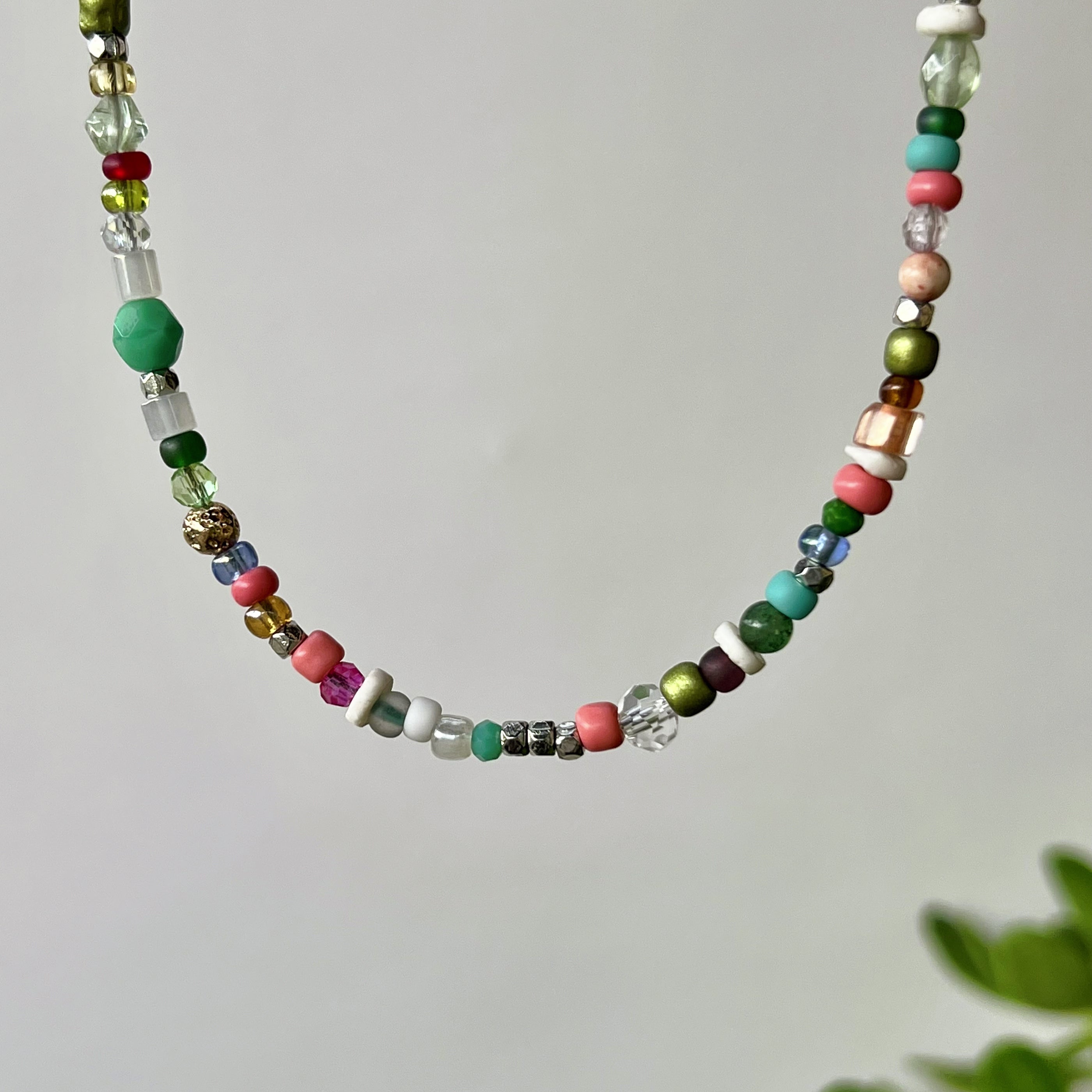 A beaded necklace with a variety of colorful glass and stone beads, including green, pink, turquoise, white, and metallic gold, hanging against a light gray background.