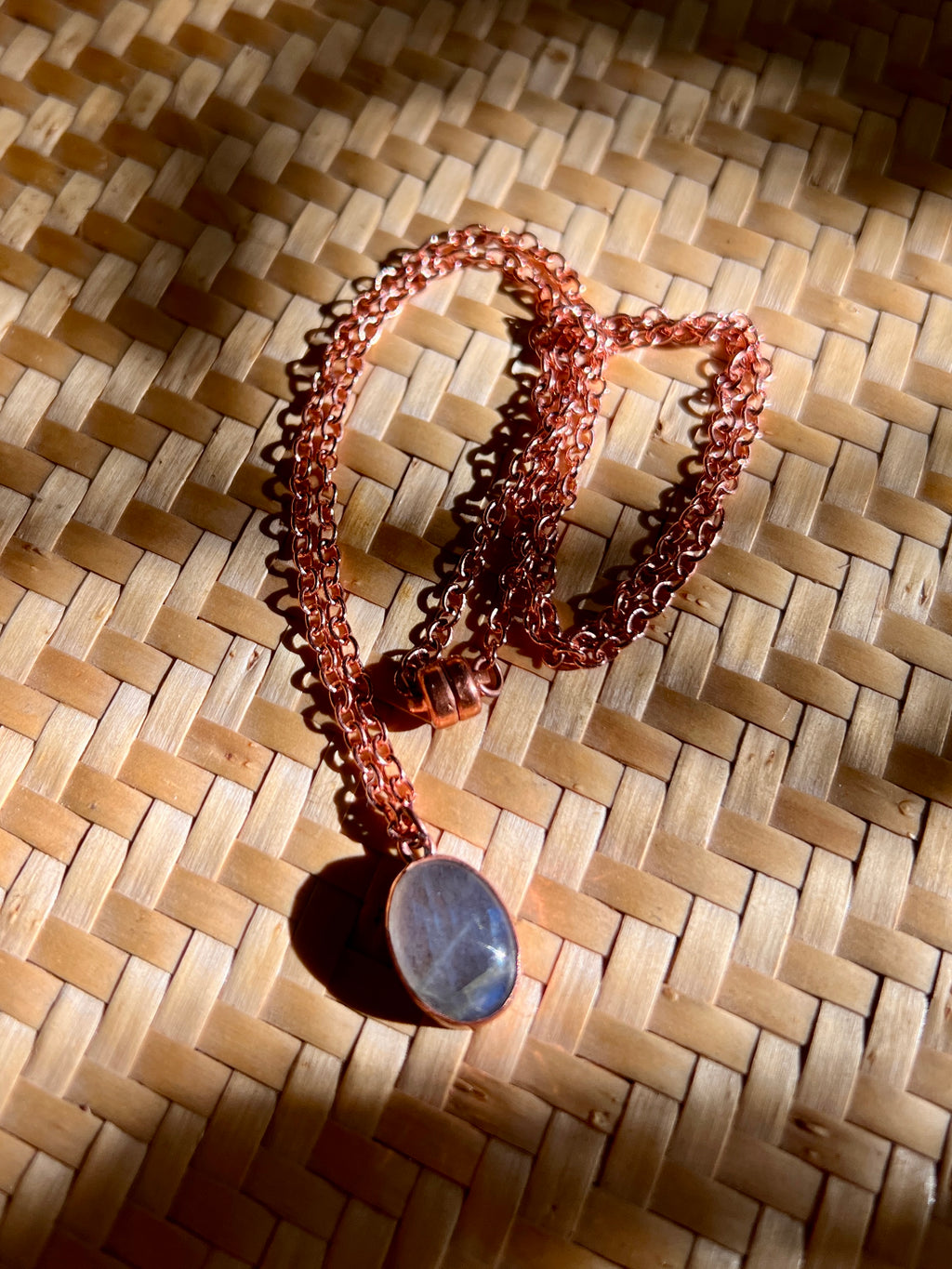 A copper chain necklace with a polished oval moonstone pendant, displayed on a woven straw mat under direct sunlight.