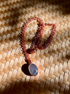A copper chain necklace with a polished oval moonstone pendant, displayed on a woven straw mat under direct sunlight.