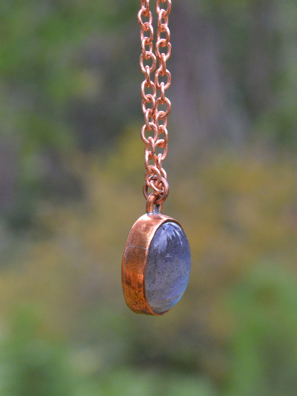 A handmade pendant necklace featuring a hammered copper bezel holding a polished slate blue oval stone, suspended from a copper chain against a blurred green and yellow outdoor background.