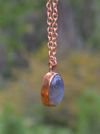 A handmade pendant necklace featuring a hammered copper bezel holding a polished slate blue oval stone, suspended from a copper chain against a blurred green and yellow outdoor background.