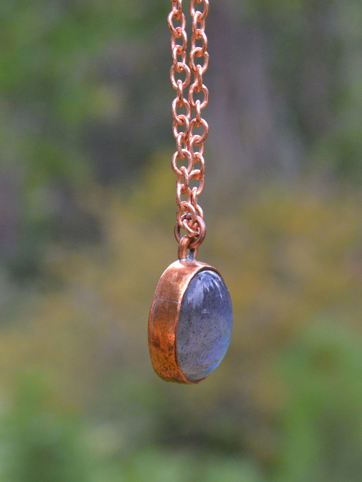 A handmade pendant necklace featuring a hammered copper bezel holding a polished slate blue oval stone, suspended from a copper chain against a blurred green and yellow outdoor background.