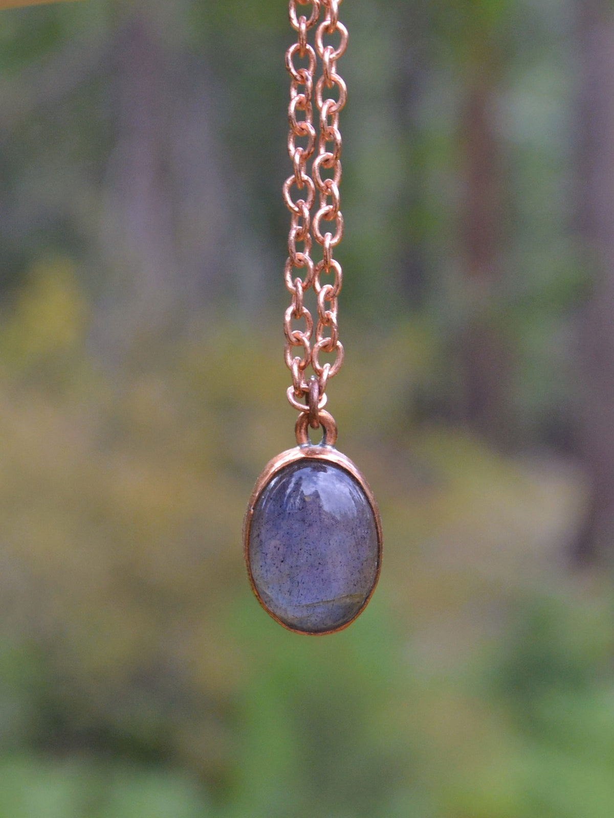 A copper chain necklace with an oval bezel containing a polished dark blue labradorite stone, shown against a blurred natural forest background.
