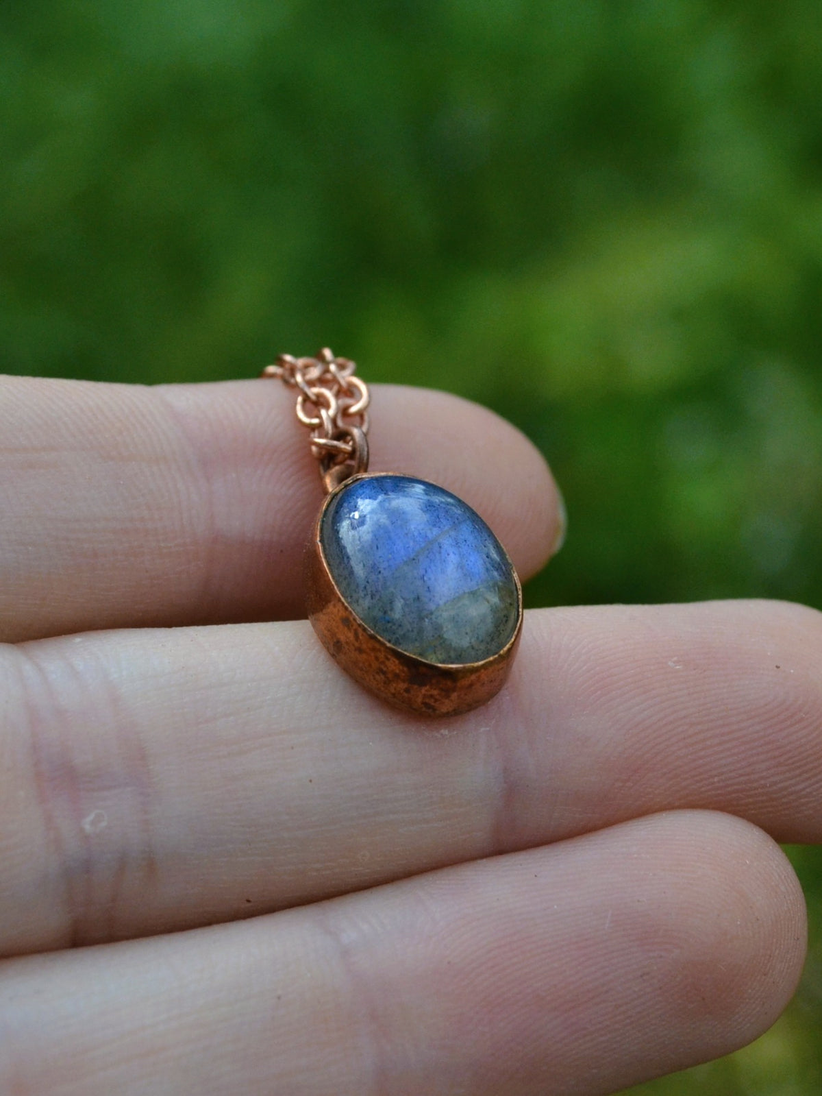 A close-up of a hand holding a small oval pendant with a blue-gray labradorite stone and a copper bezel and chain, against a blurred green outdoor background.