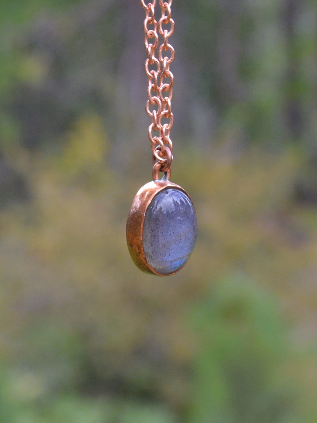 A close-up of a copper chain necklace with an oval pendant featuring a blue-gray stone set in a copper bezel, photographed against a blurred outdoor background.