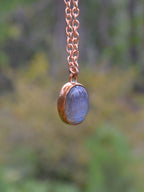 A close-up of a copper chain necklace with an oval pendant featuring a blue-gray stone set in a copper bezel, photographed against a blurred outdoor background.