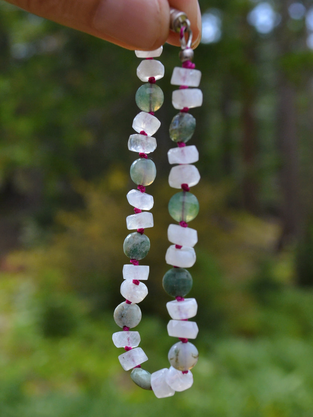 A beaded bracelet made of alternating translucent white and green stone beads strung on a magenta cord, held up by a hand against a blurred outdoor forest background.