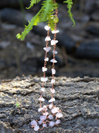 A copper chain necklace featuring natural light pink quartz stones, hanging outdoors against a dark textured log with green fern leaves in the background.
