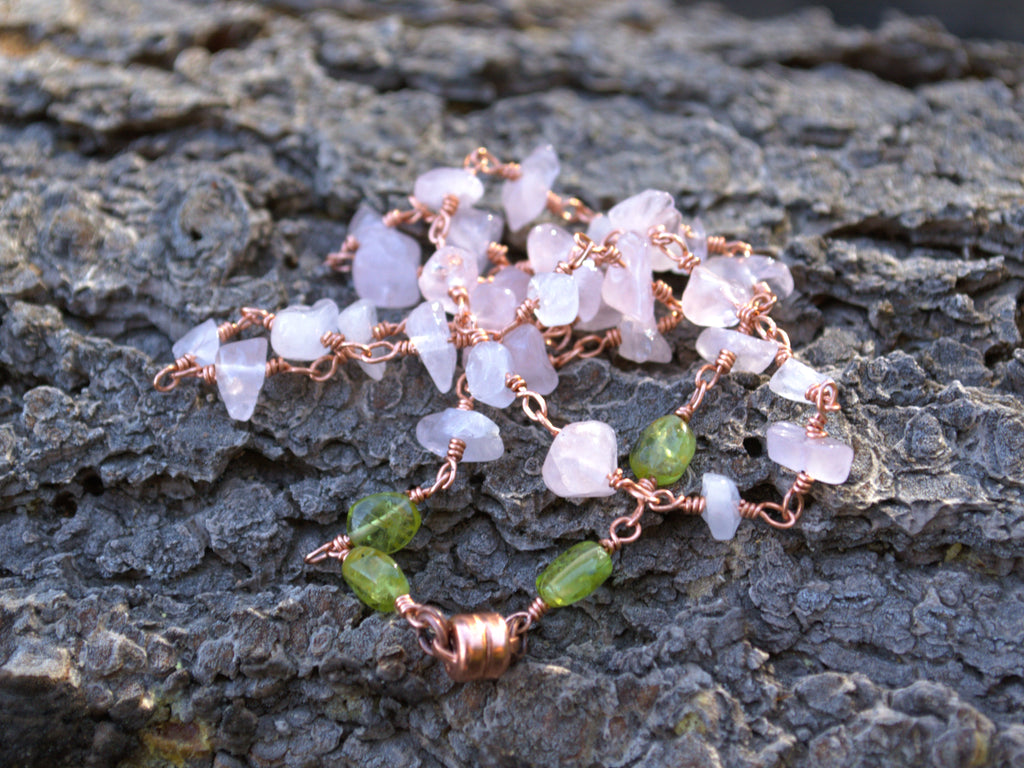 A handmade wire-wrapped gemstone necklace featuring pale pink quartz and green beads, displayed on a textured dark gray bark surface.