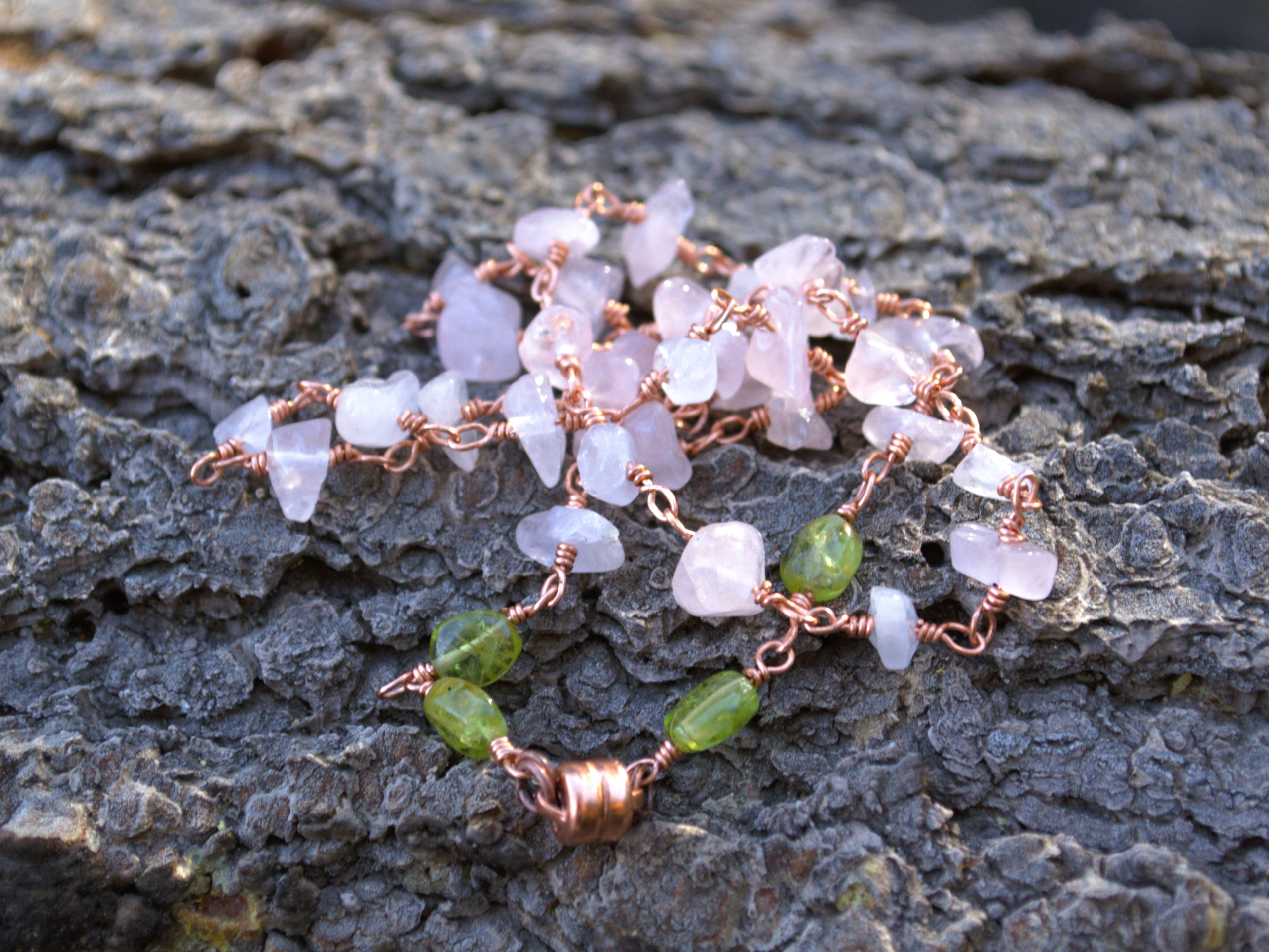 A handmade wire-wrapped gemstone necklace featuring pale pink quartz and green beads, displayed on a textured dark gray bark surface.