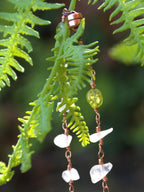 A close-up of a handmade copper wire dangle earring with a small green faceted bead and clear crystal chips, hanging against a background of green fern leaves.