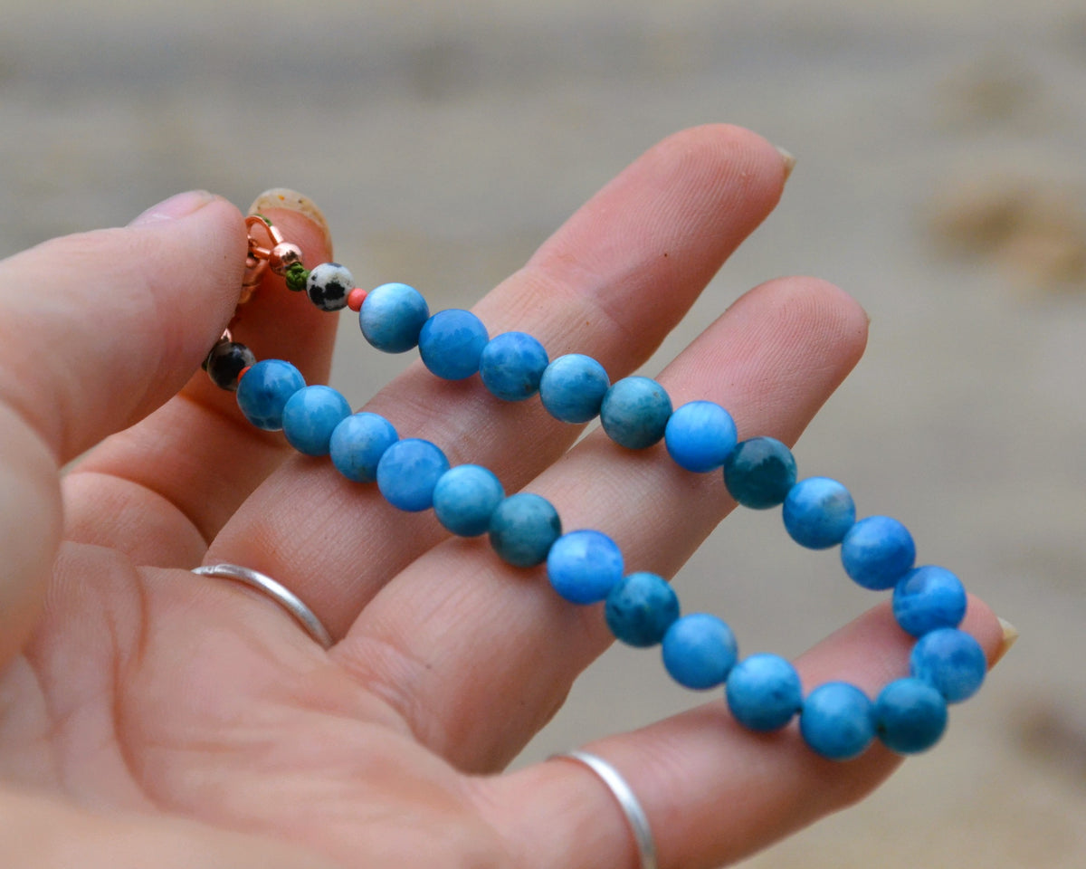 A close-up of a hand holding a beaded bracelet made of round blue marbled beads, with tiny accent beads near the clasp, against a blurred sandy background.