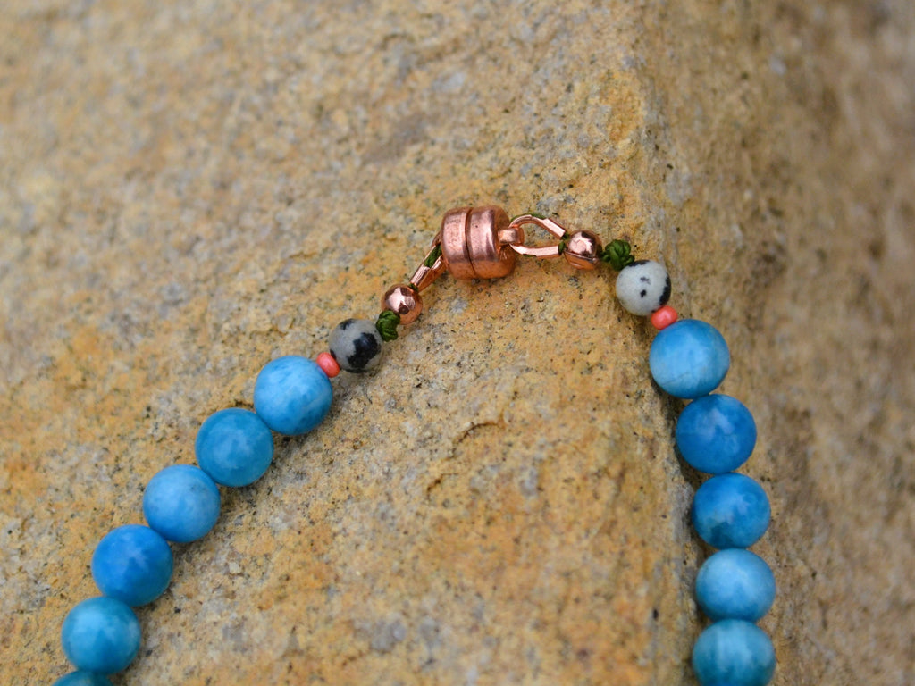 A close-up of a beaded bracelet with round blue beads, a copper clasp, and small accent beads, resting on a textured tan rock surface.
