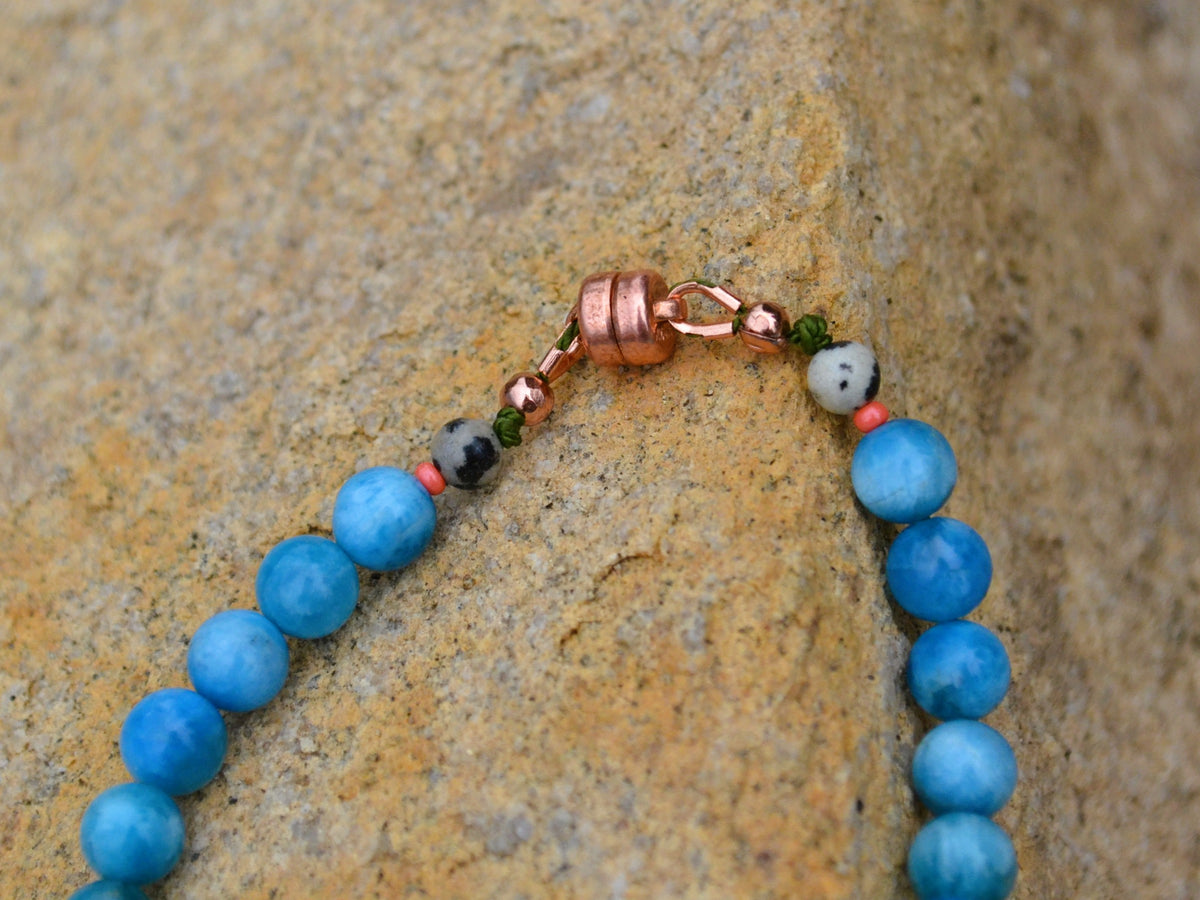 A close-up of a beaded bracelet with round blue beads, a copper clasp, and small accent beads, resting on a textured tan rock surface.