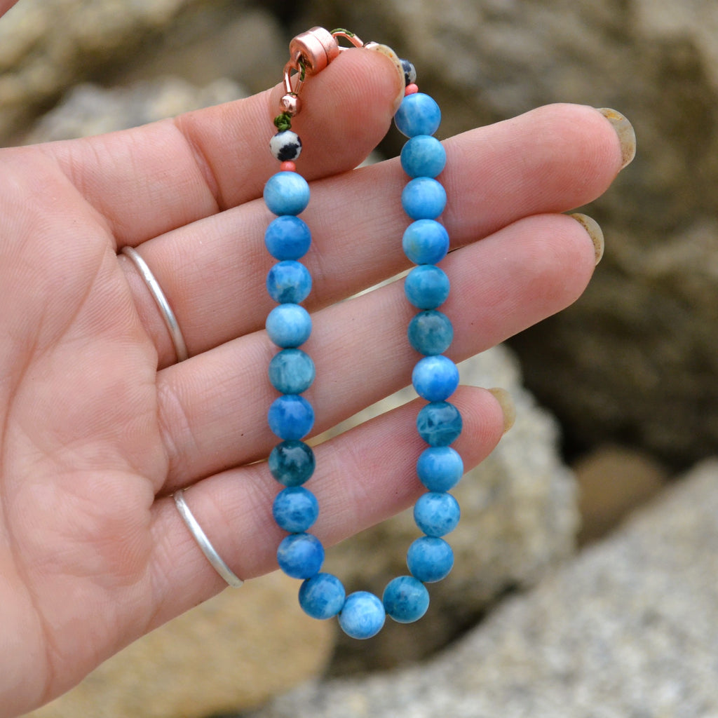 A beaded bracelet made of round blue apatite gemstone beads with a copper clasp, held in a person's hand against a blurred rocky outdoor background.