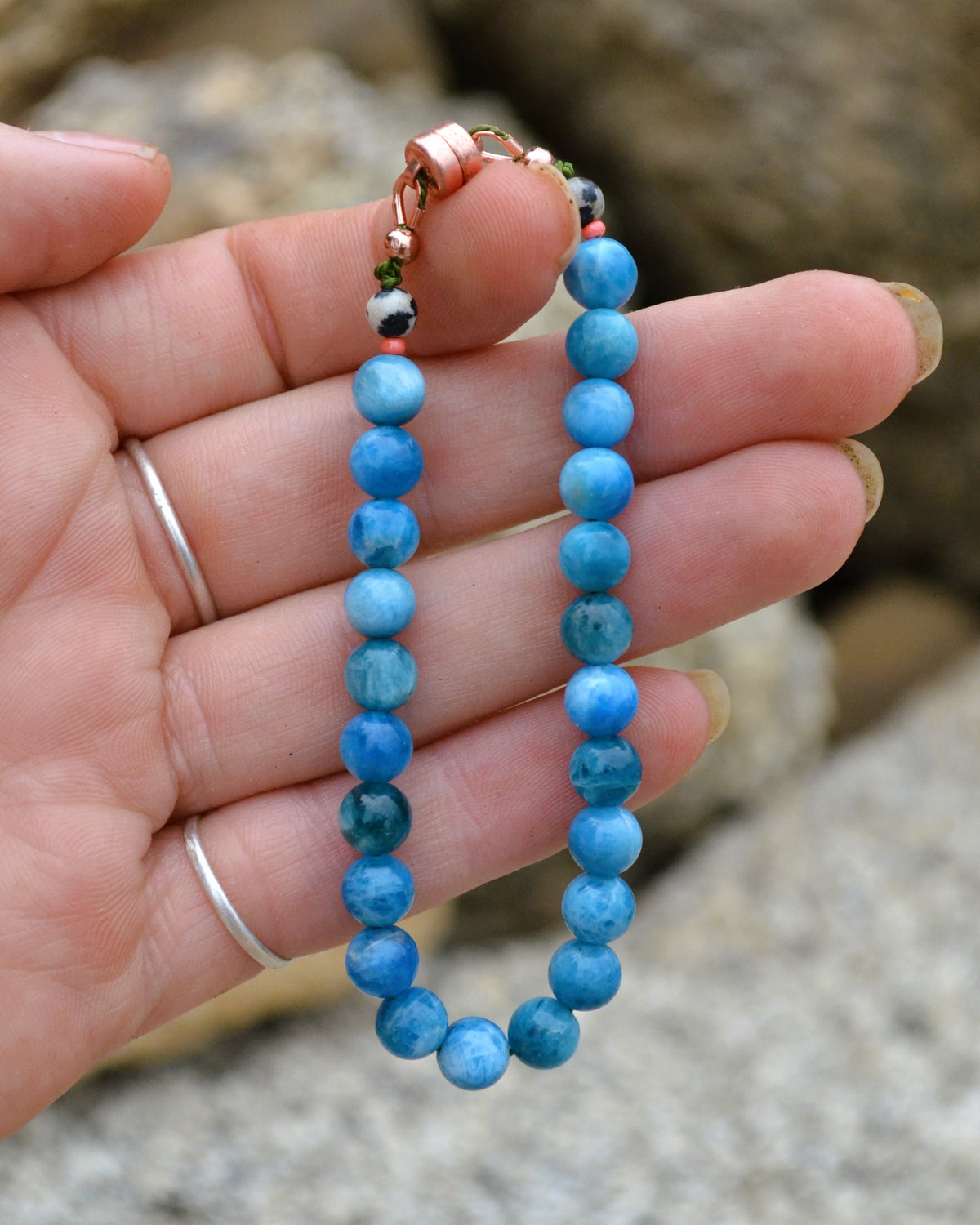 A beaded bracelet made of round light blue marbled beads with a copper clasp, held in a person's hand against a blurred outdoor rocky background.