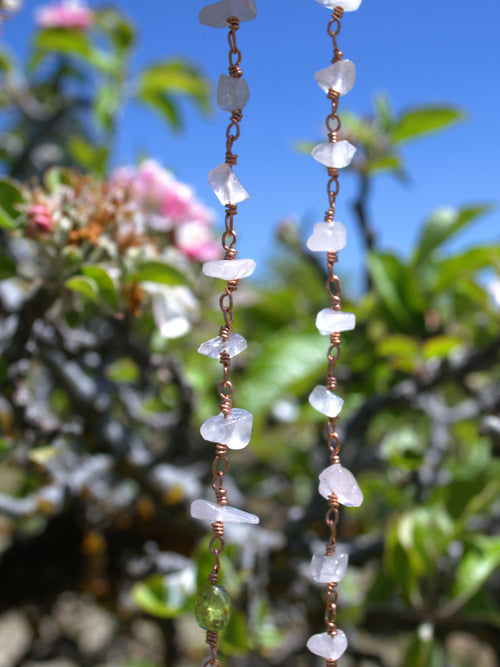 A close-up of two strands of a handmade beaded necklace made of irregular white quartz stones and copper wire, with a small green bead accent, set against a blurred background of green foliage and blue sky.