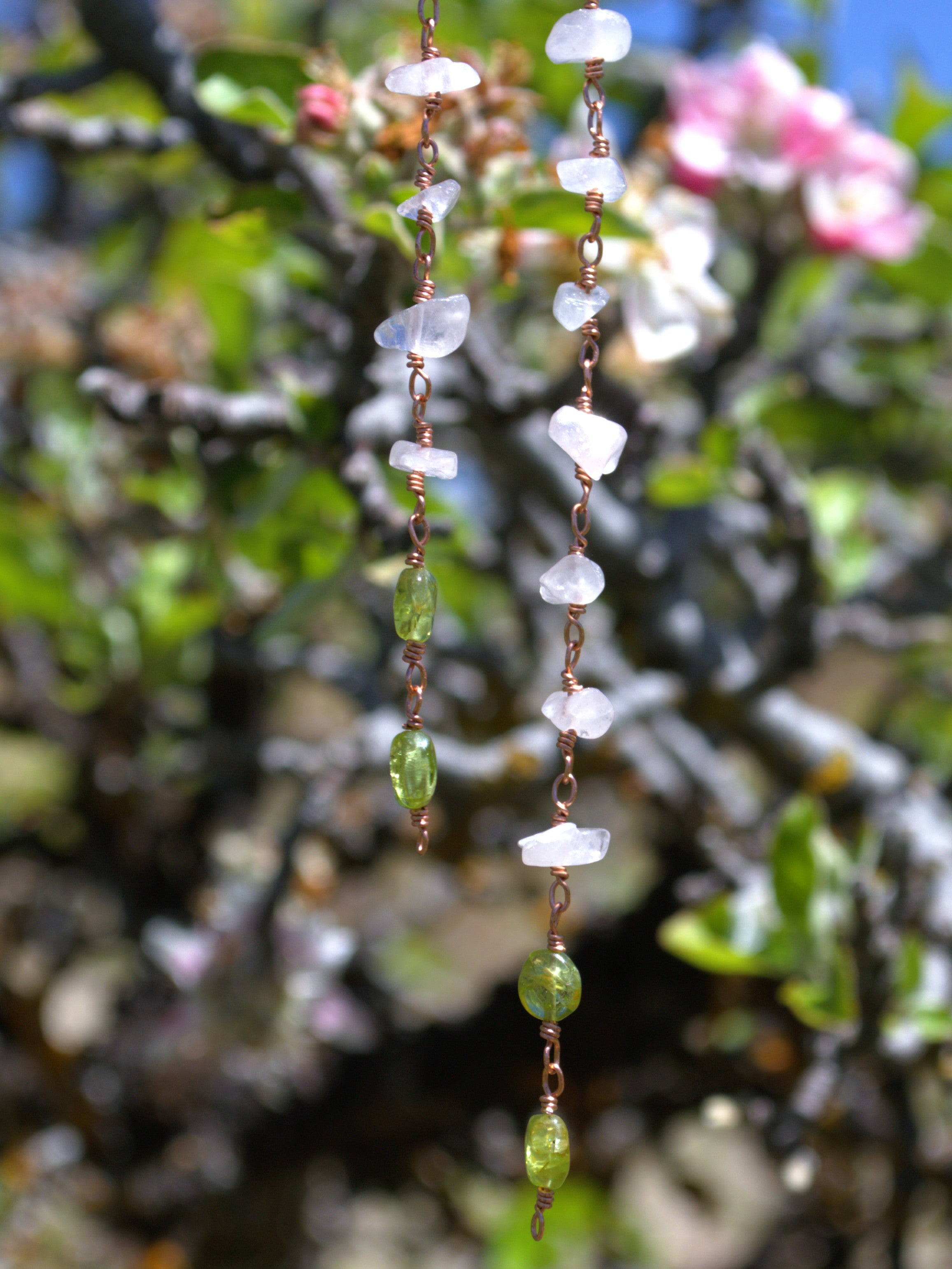 A close-up of two hanging strands of handmade jewelry featuring white translucent stone chips, green faceted beads, and copper wire spacers, set against a blurred background of green leaves and pink flowers.