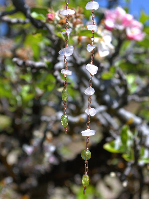 A close-up of two hanging strands of handmade jewelry featuring white translucent stone chips, green faceted beads, and copper wire spacers, set against a blurred background of green leaves and pink flowers.