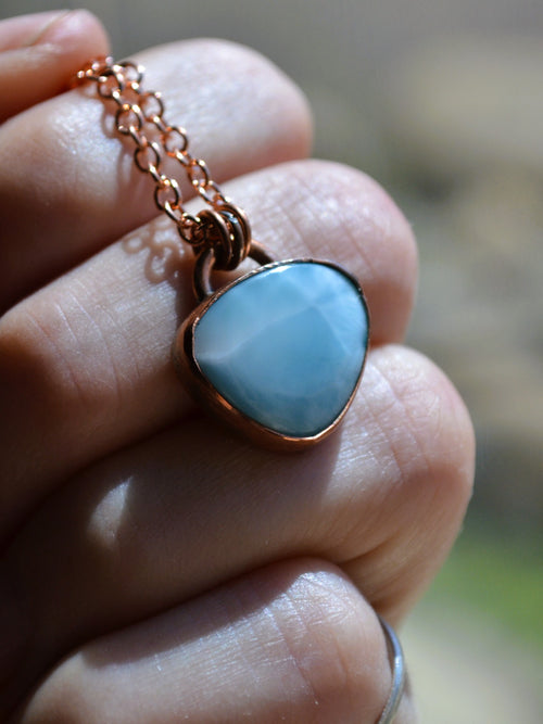 A close-up of a hand holding a pendant necklace. The pendant features a light blue marbled stone set in a copper bezel, suspended from a matching copper chain.