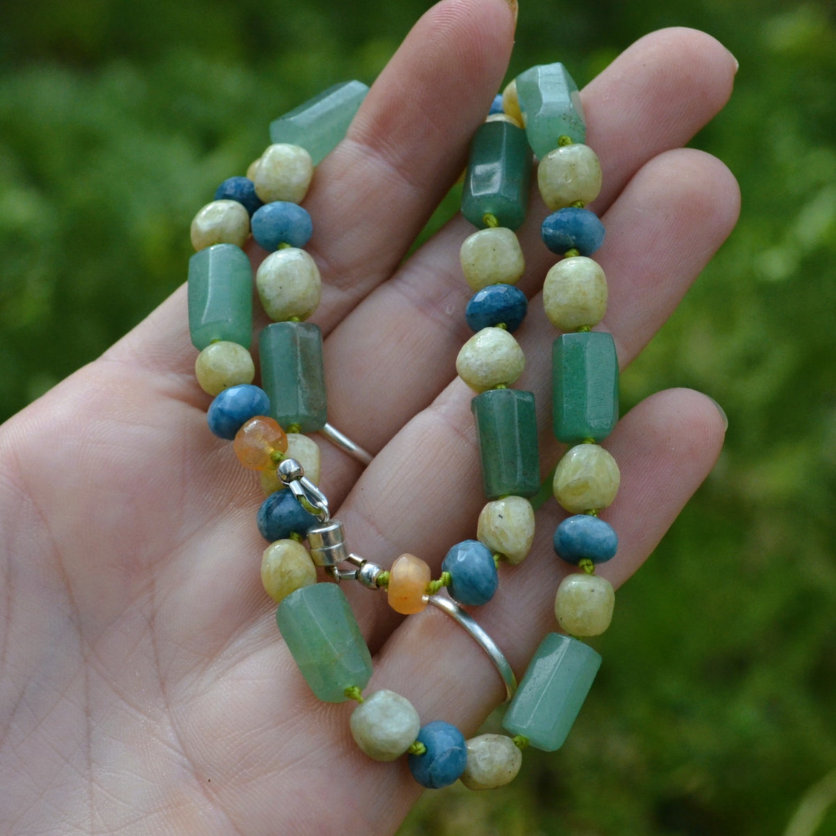 A beaded strand necklace with green, blue, and pale yellow beads, held in a person's hand against a blurred green outdoor background.