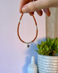 Hand holding an orange beaded bracelet with a silver heart charm against a blurred indoor background