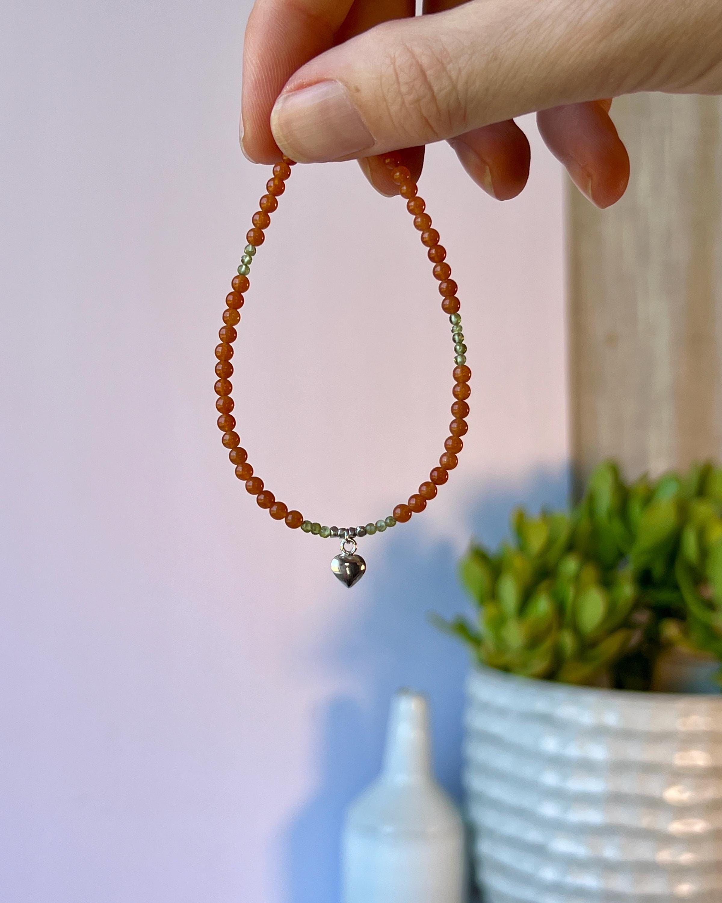 Hand holding an orange beaded bracelet with a silver heart charm against a blurred indoor background