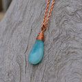 A teardrop-shaped aqua blue stone pendant necklace wrapped in copper wire, hanging from a copper chain against a textured gray wood background.