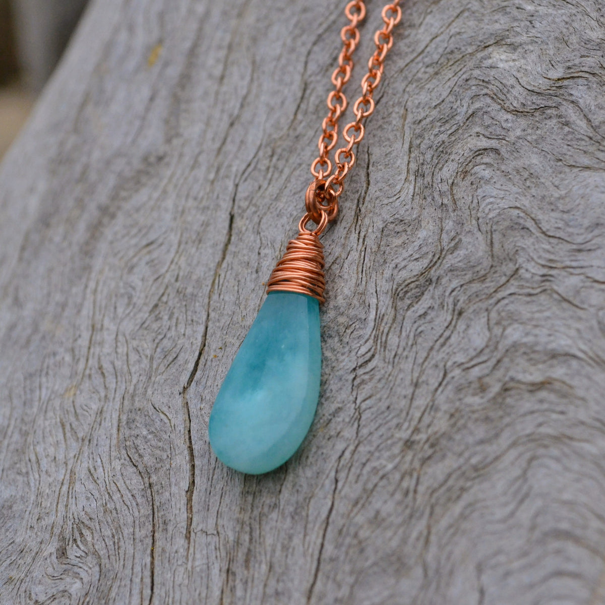 A teardrop-shaped aqua blue stone pendant necklace wrapped in copper wire, hanging from a copper chain against a textured gray wood background.
